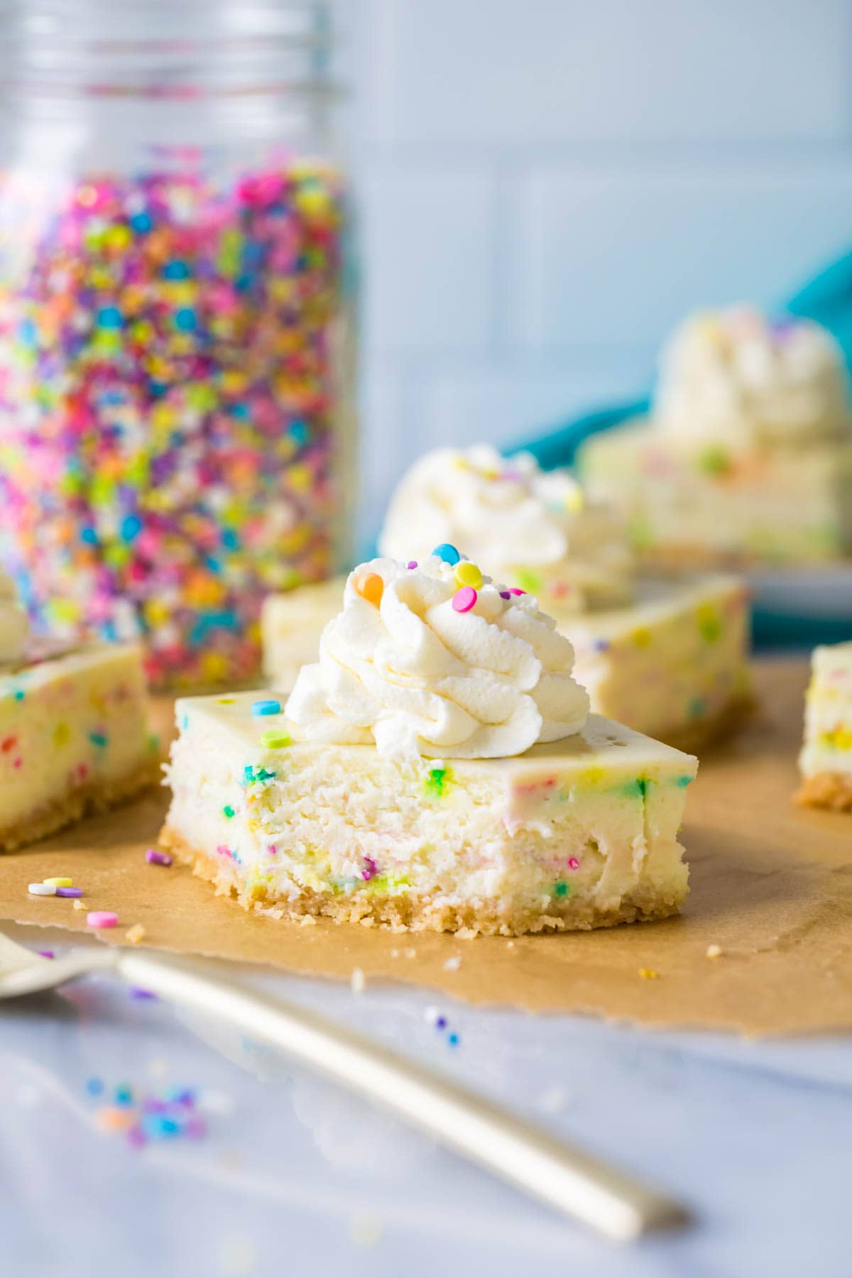 sprinkle-studded cheesecake bar with a bite out of it; gold fork in foreground and jar of sprinkles in background