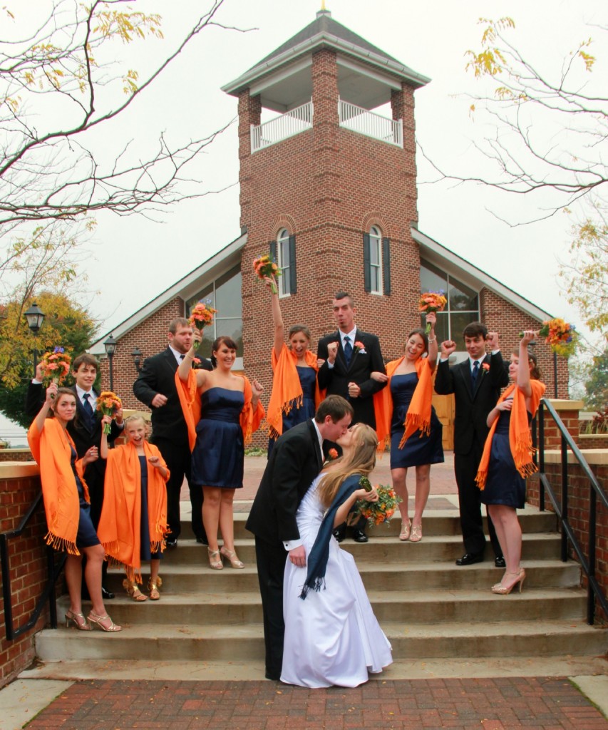 bride and groom kiss in front of church, bridal party celebrates in background