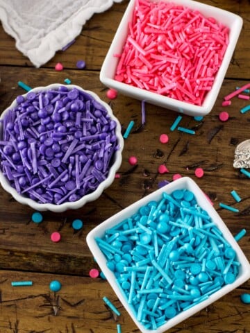 Three bowls: one filled with blue sprinkles, one with pink sprinkles, one with purple sprinkles