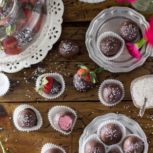 Overhead of strawberry buttercream candies arranged on a silver plate and wood surface, surrounded by chocolate covered strawberries