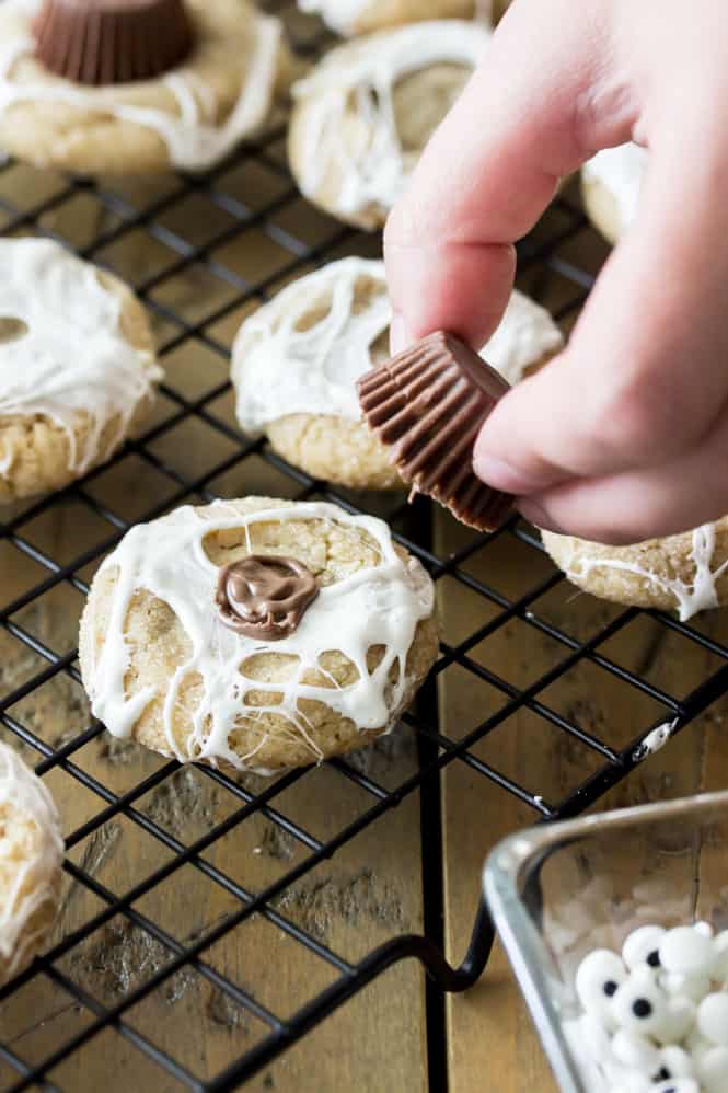 Hand lowering mini reeses cup on to spiderwebbed cookie; cookie is topped with chocolate dollop to adhere the reeses cup