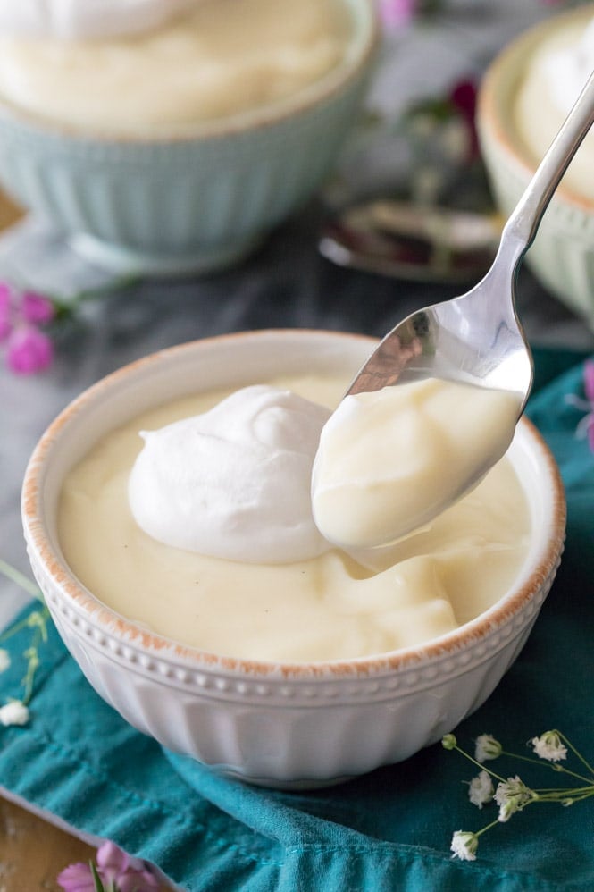 Spoon with vanilla pudding on it, bowl of vanilla pudding topped with whipped cream in the background