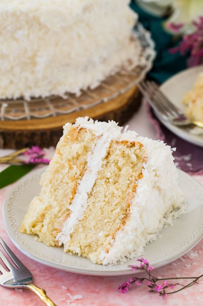 Slice of layered coconut cake on white plate on pink backdrop