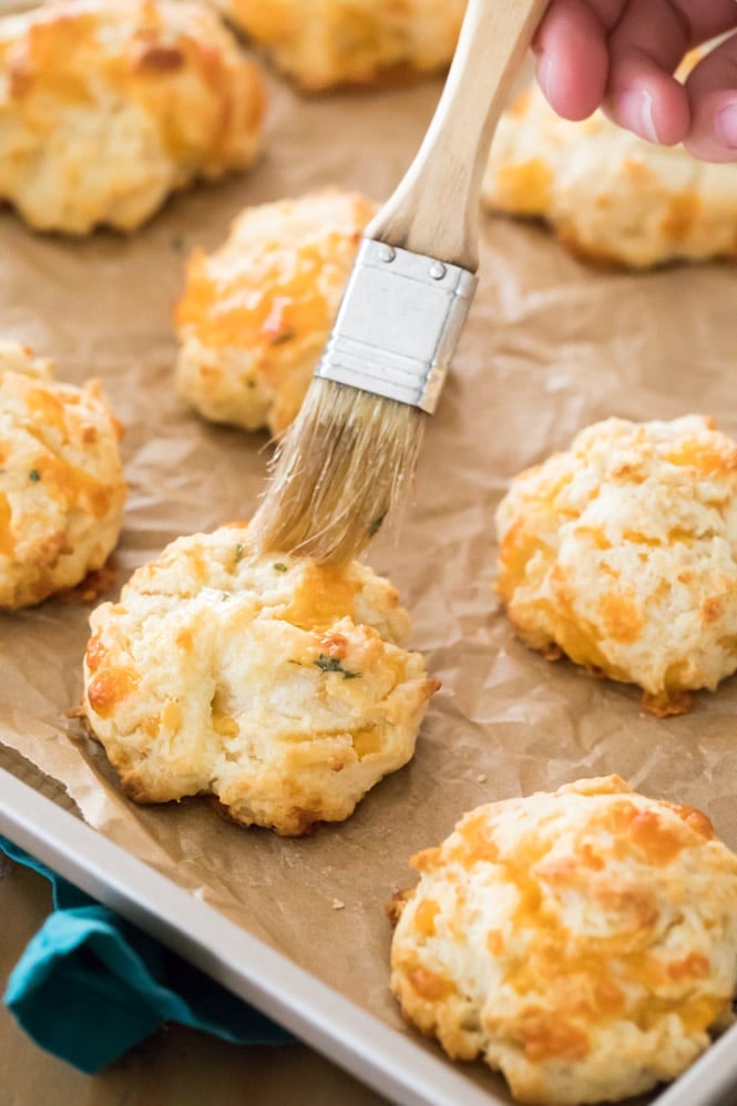 Brushing butter over a freshly baked garlic cheese drop biscuit on a baking sheet