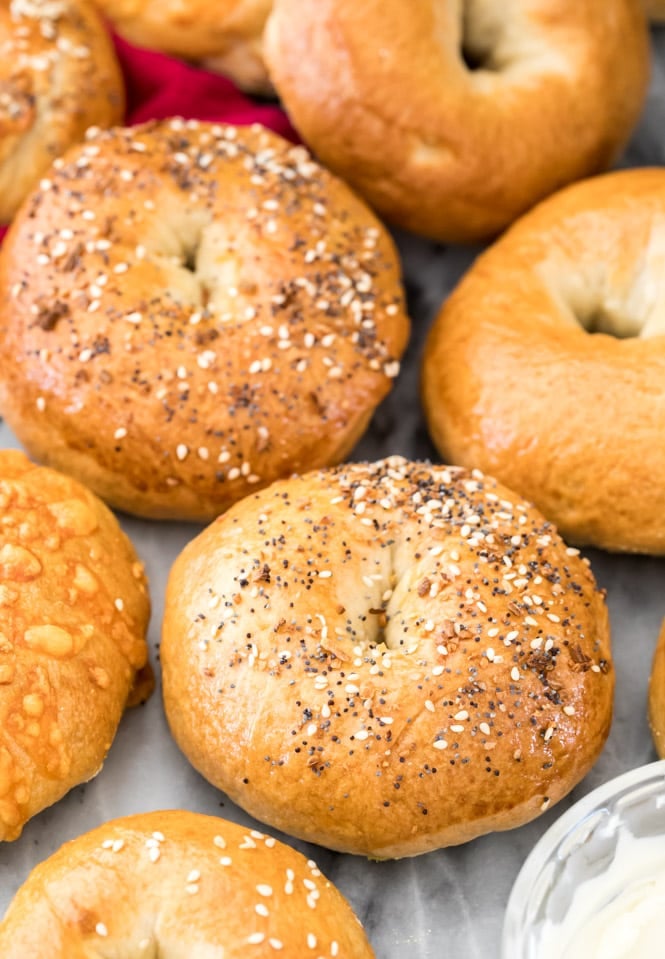a variety of bagels on marble next to a bowl of cream cheese