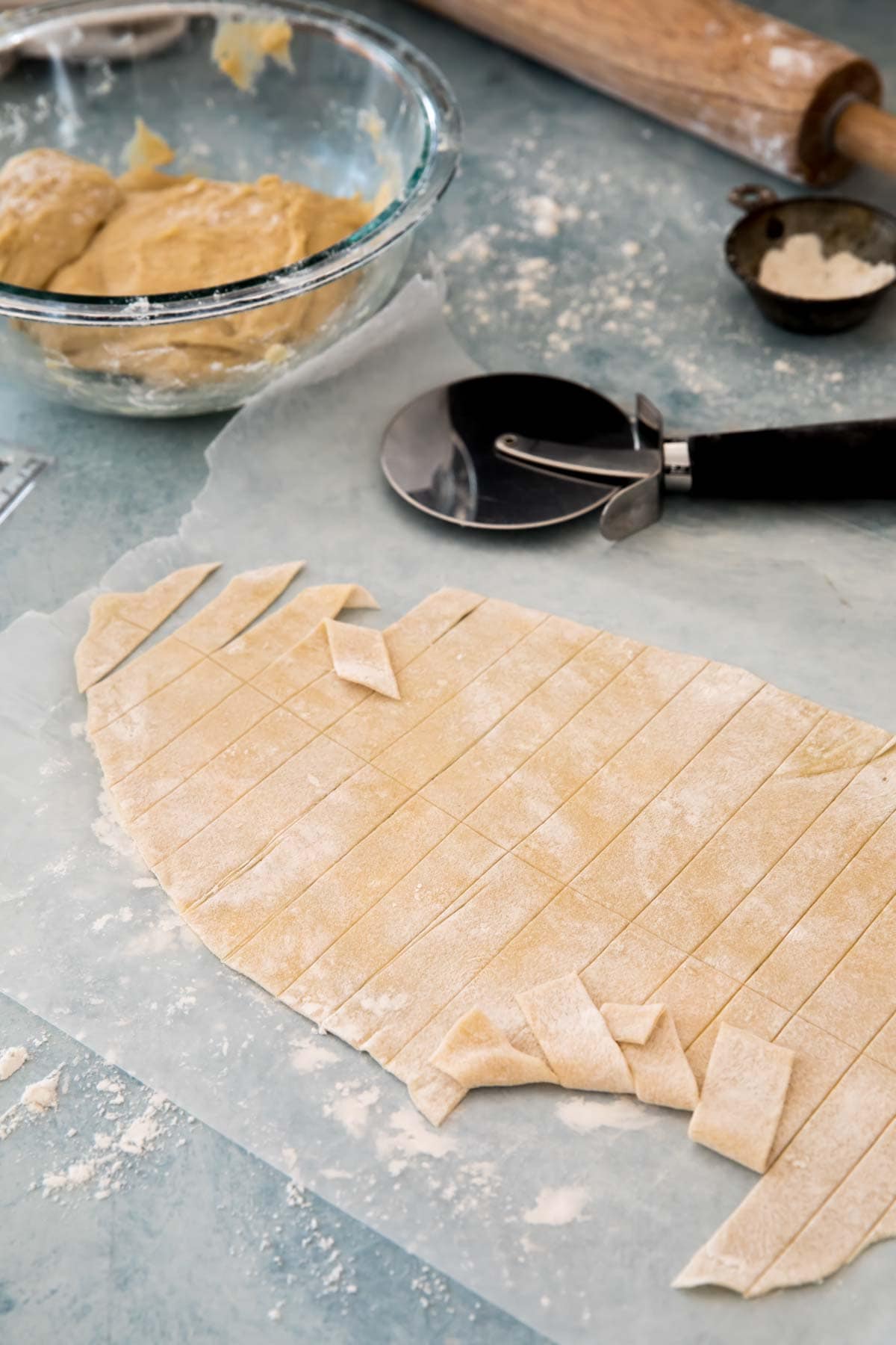 Dough rolled paper thin and cut into strips. pizza cutter, rolling pin, flour, and glass bowl with more dough in background