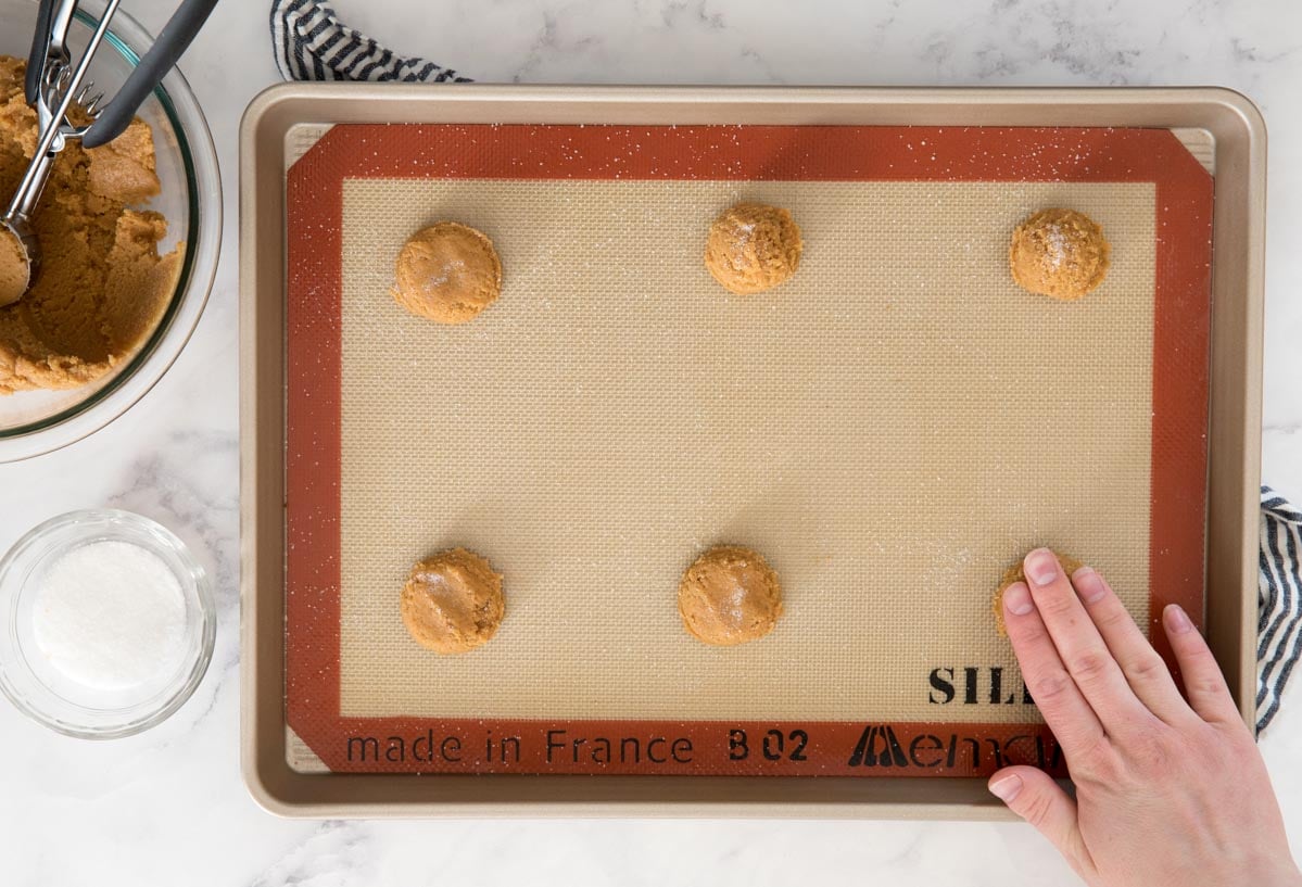 Cookie dough balls on a baking sheet being flattened with clean fingers