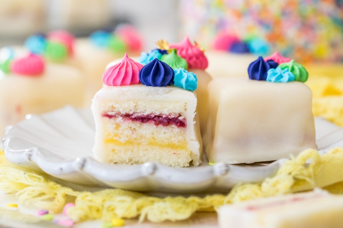 cross-section of petit four on white plate with frosting showing cake, lemon curd, and raspberry layers