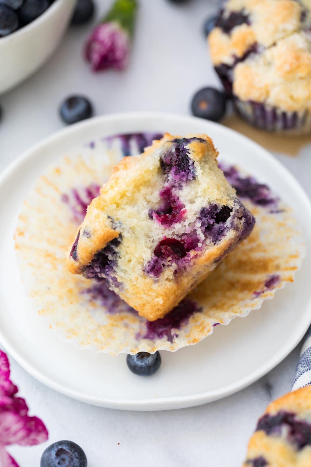 blueberry muffin on wrapper on white plate with a bite taken out of it to show fluffy interior