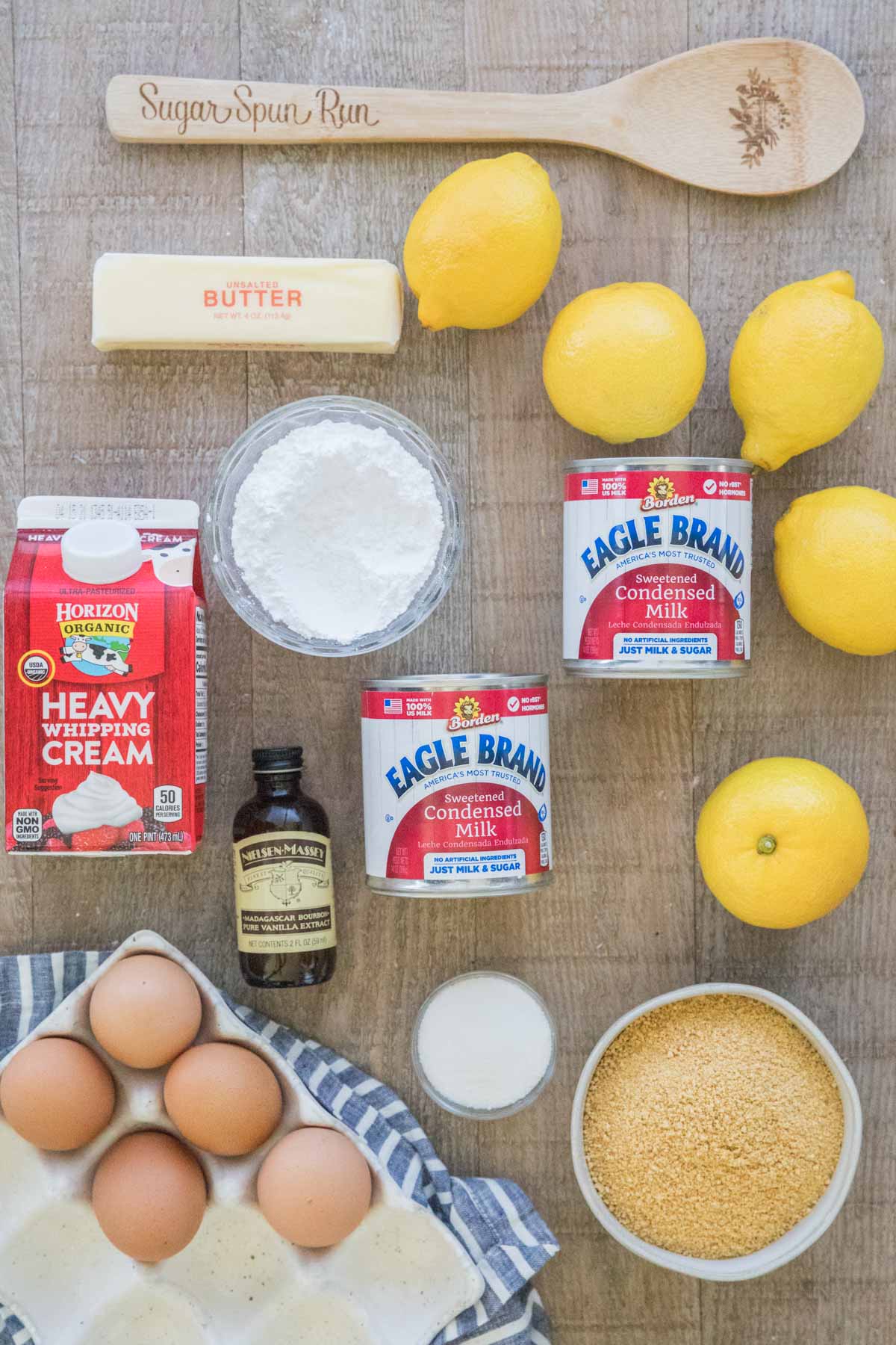 Wooden spoon, butter, lemons, powdered sugar, sweetened condensed milk, heavy cream, vanilla extract, eggs, granulated sugar, and graham cracker crumbs.