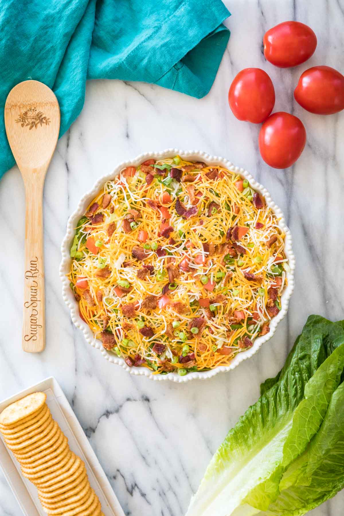 Overhead of blt dip in round white dish surrounded by vegetables, crackers, a teal towel, and a wooden spoon