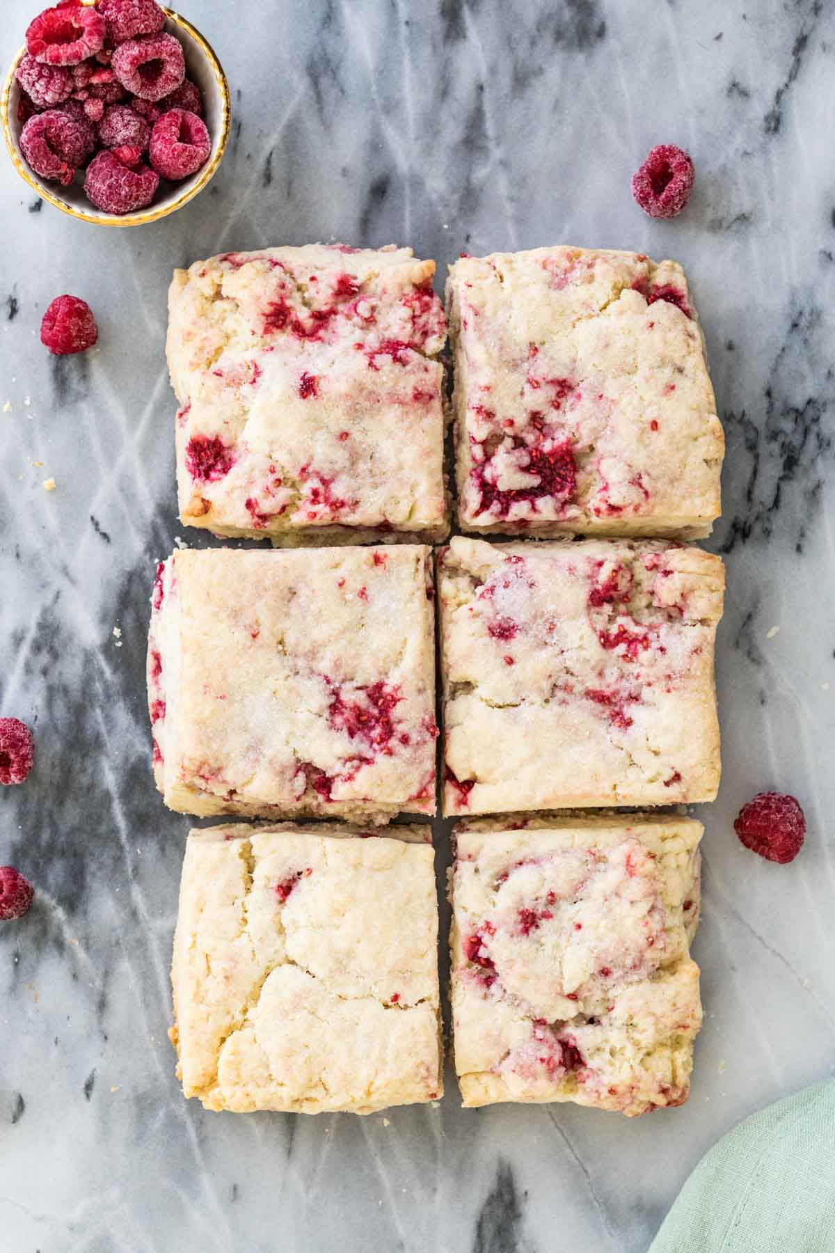 Overhead shot of raspberry scones on gray marble next to a bowl of raspberries in the top left corner