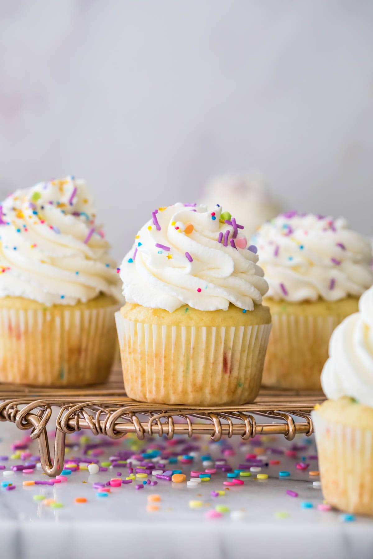 Closeup of vanilla sprinkle cupcake with whipped cream frosting and sprinkles, surrounded by more frosted cupcakes