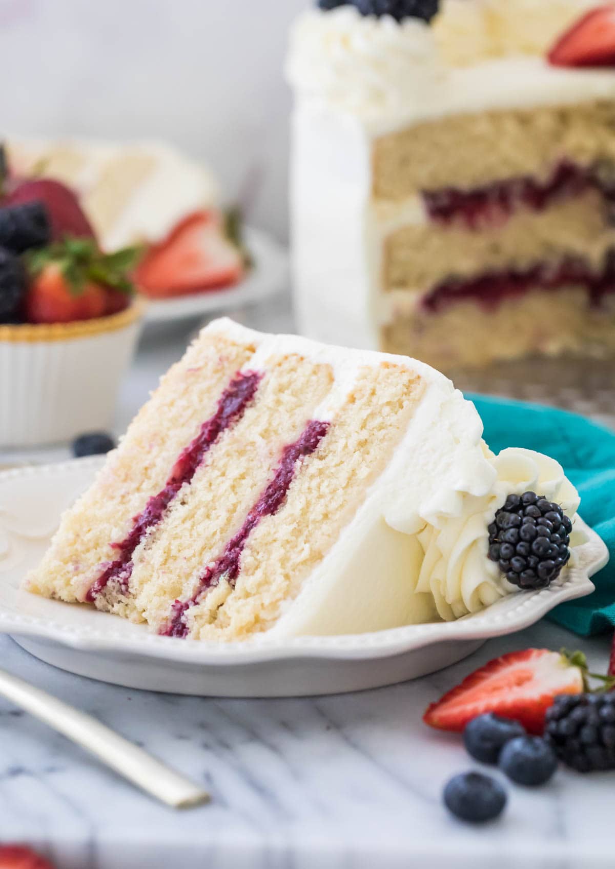 Three layer chantilly cake with berry filling on white plate surrounded by fruit and more cake