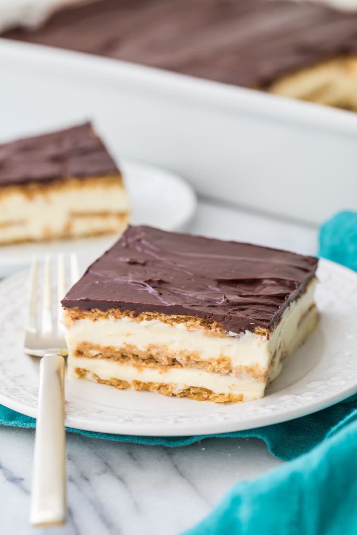 Slice of eclair cake showing two layers of pastry cream on a white plate with a teal towel beneath the plate