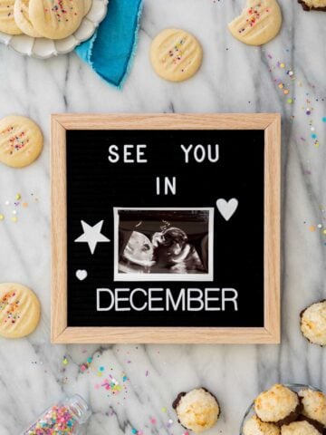 Felt board that reads: "See you in December" with an ultrasound photo in the center. Board is on white marble surrounded by cookies and sprinkles