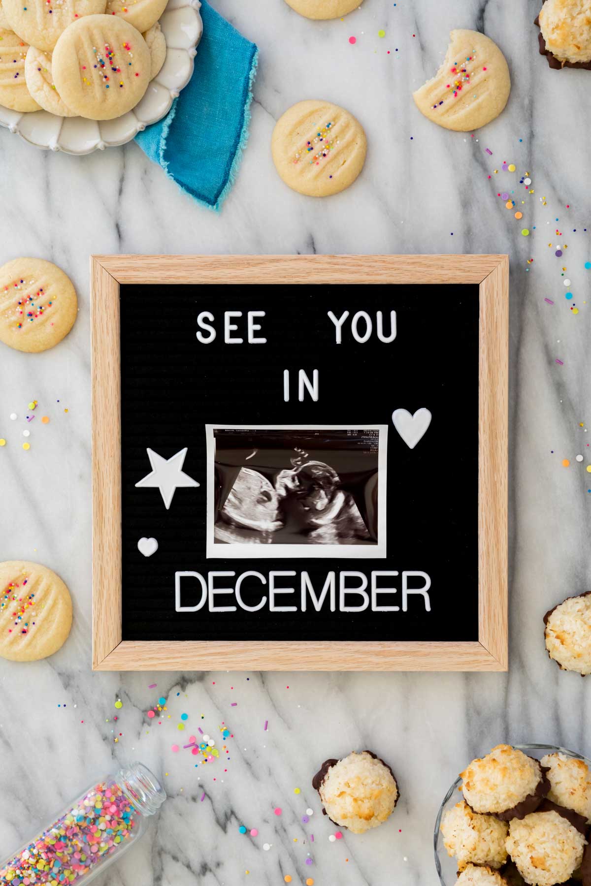 Felt board that reads: "See you in December" with an ultrasound photo in the center. Board is on white marble surrounded by cookies and sprinkles