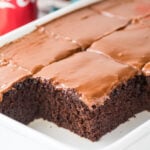 Thick squares of coca cola cake with shiny brown frosting in white cake pan