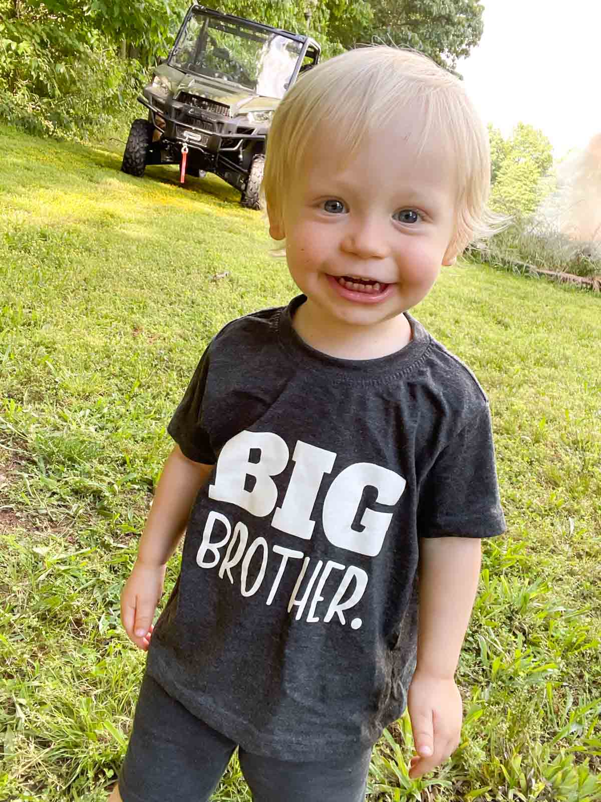 Toddler boy in a grey shirt that reads "BIG BROTHER". Photo taken outside, side by side in background