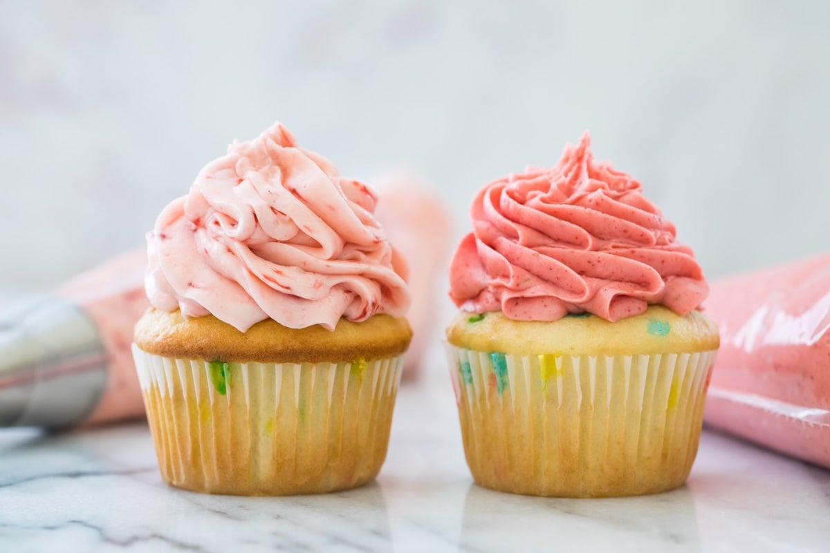 Two yellow sprinkle cupcakes, the left with a pale pink flecked strawberry frosting and the right with a more vibrant pink frosting.