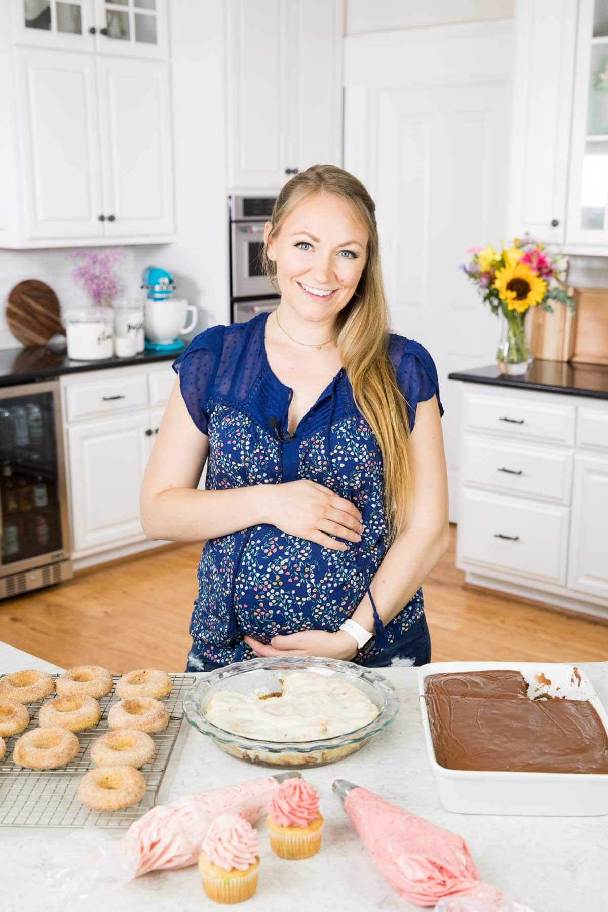 The author in a blue shirt holding her pregnant belly in the kitchen. On the counter in front of her is an array of desserts