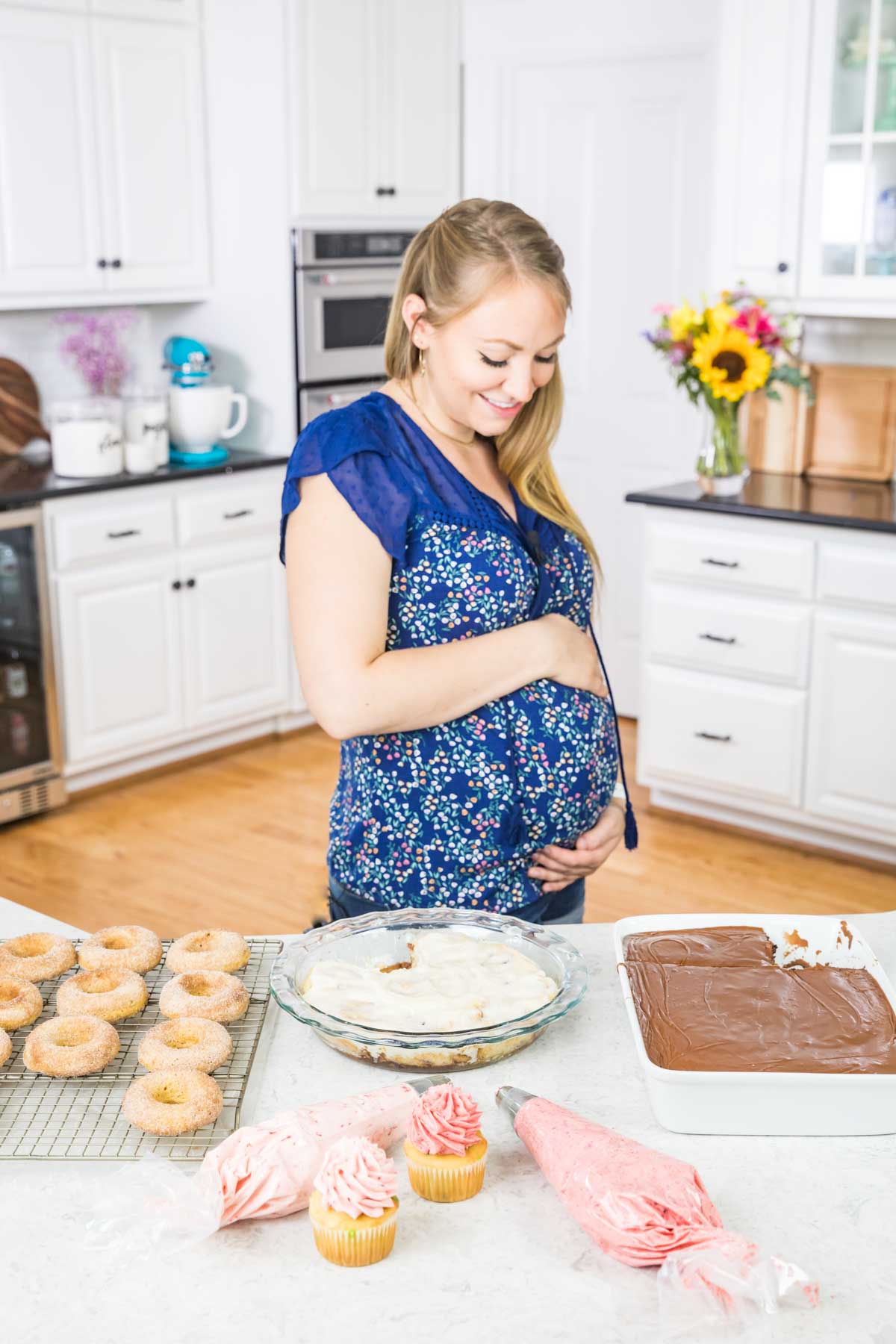 The author in a blue shirt holding her pregnant belly in the kitchen. On the counter in front of her is an array of desserts