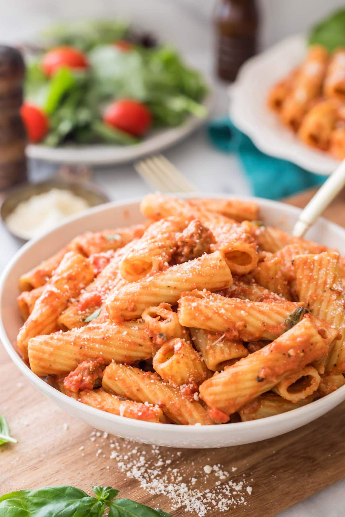 large white serving bowl of rigatoni with salad in the background