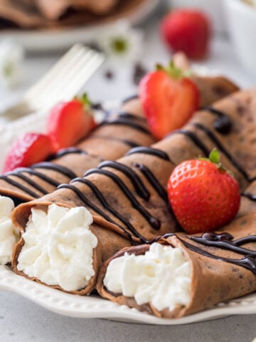 three chocolate crepes rolled and filled with whipped creme topped with chocolate sauce and strawberries