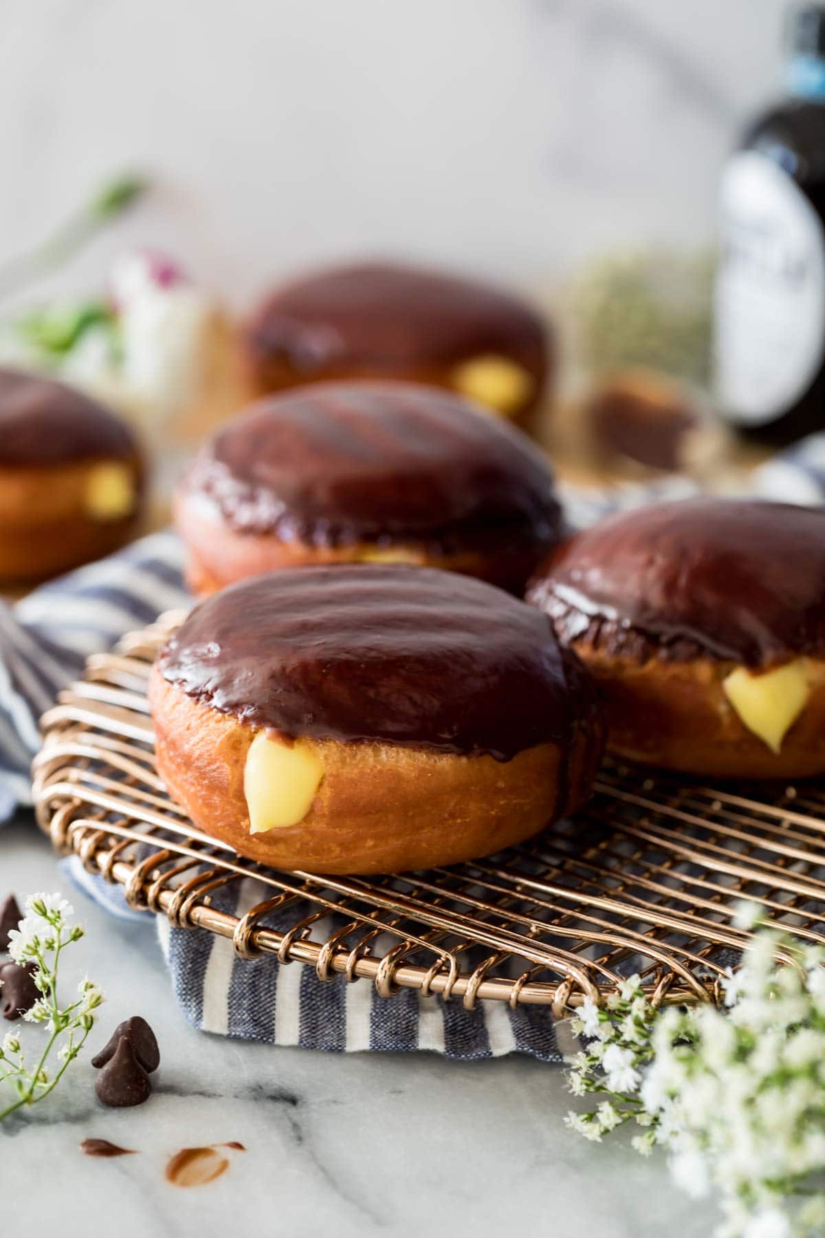 Three Boston cream donuts covered in chocolate ganache and filled with pastry cream filling on a metal cooling rack, with more donuts in the background