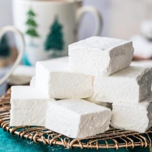 square-shaped homemade marshmallows stacked on a gold metal cooling rack with mugs of hot chocolate in the background