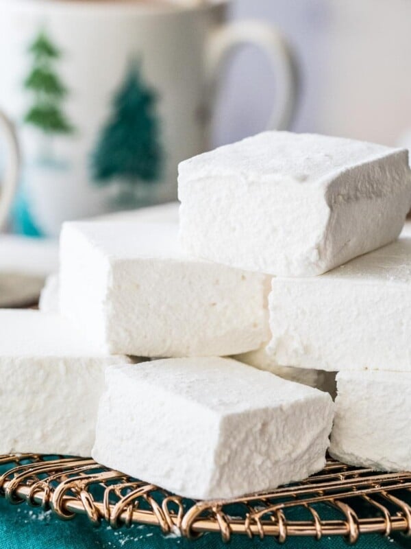 square-shaped homemade marshmallows stacked on a gold metal cooling rack with mugs of hot chocolate in the background