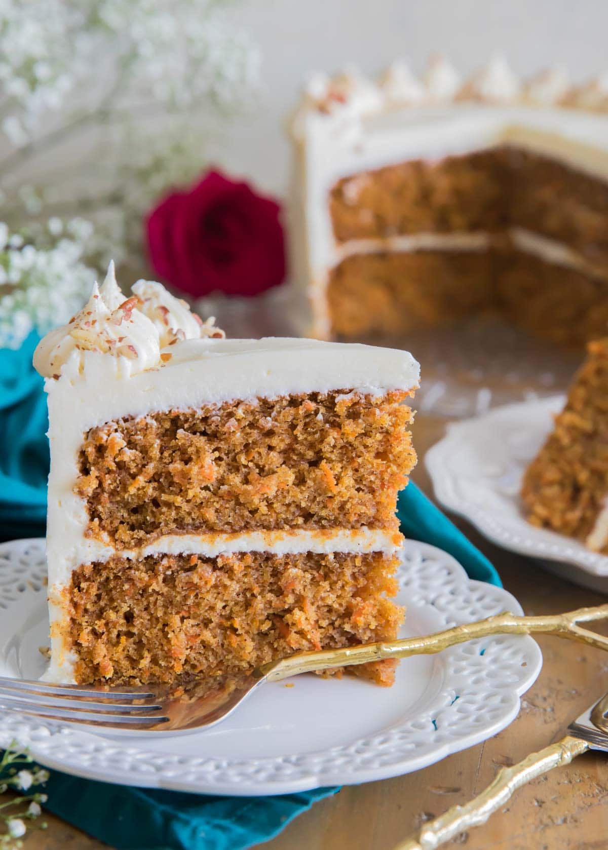 a slice of frosted carrot cake on a white plate with the rest of the cake in the background