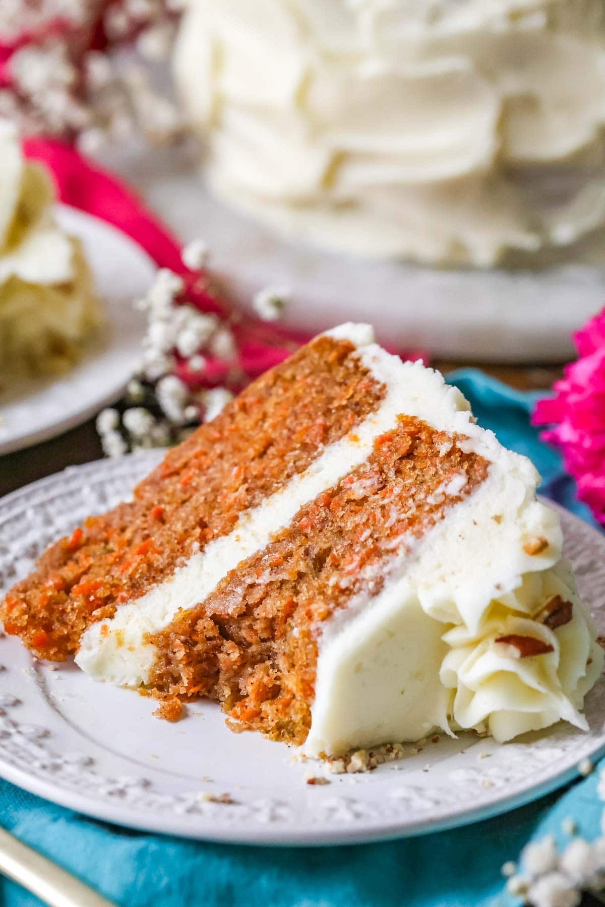This is an incredibly moist, perfect, BEST Carrot Cake recipe, hands down! #carrotcake #bestcake #baking #recipe #classicrecipe A slice of the best carrot cake recipe on a white plate with white flowers in the background. A bite missing from the slice.