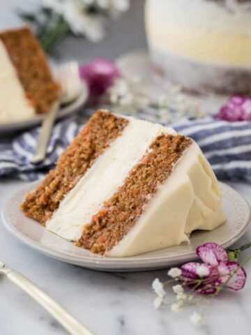 two thick slices of carrot cake cheesecake on white plates with the remaining cake in the background