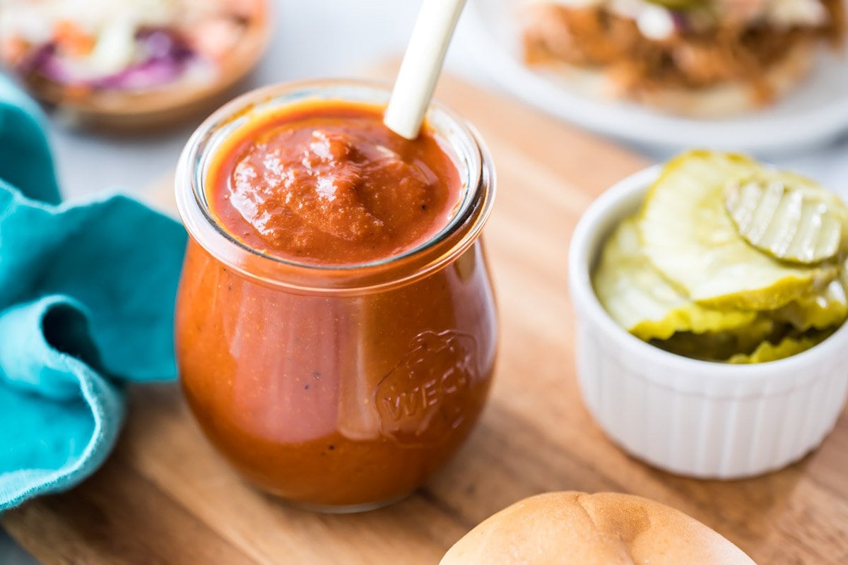 small jar of barbecue sauce on a wood cutting board with pickles, coleslaw, and pulled pork in the background