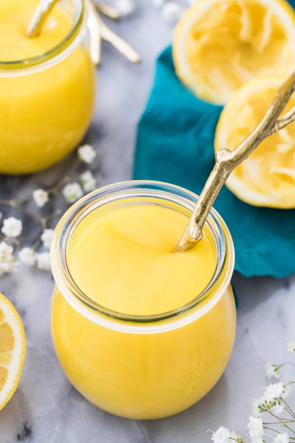 glass jar full of homemade lemon curd on a gray marble surface surrounded by lemon halves and small white baby's breath flowers