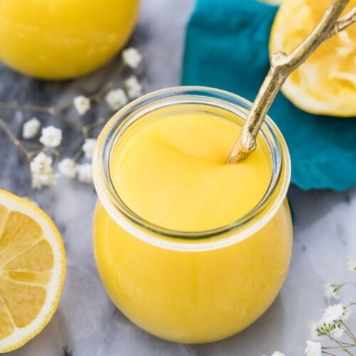 glass jar full of homemade lemon curd on a gray marble surface surrounded by lemon halves and small white baby's breath flowers