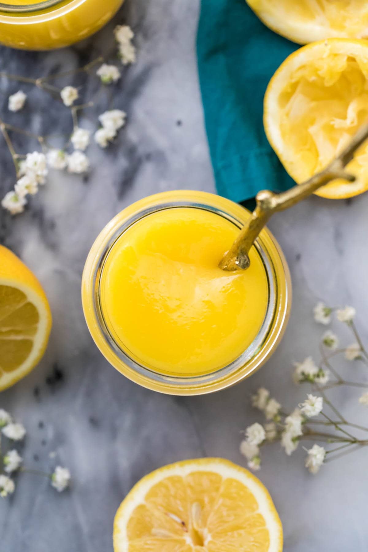 overhead view of a glass jar filled with yellow curd on a gray marble surface scattered with lemon halves and white baby's breath flowers