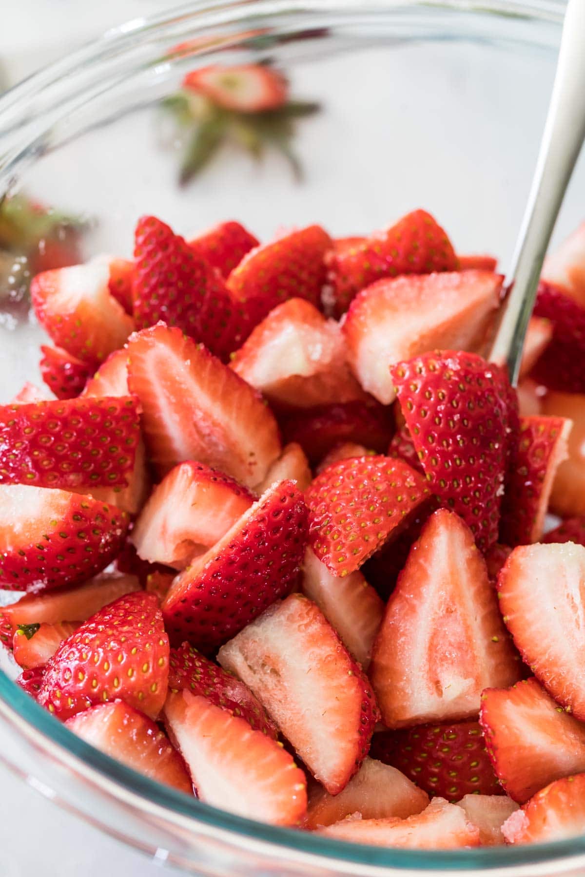 metal spoon stirring quartered strawberries in a clear bowl