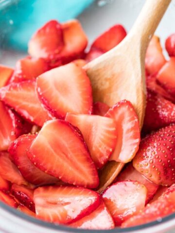 wooden spoon stirring sliced macerated strawberries in a clear bowl