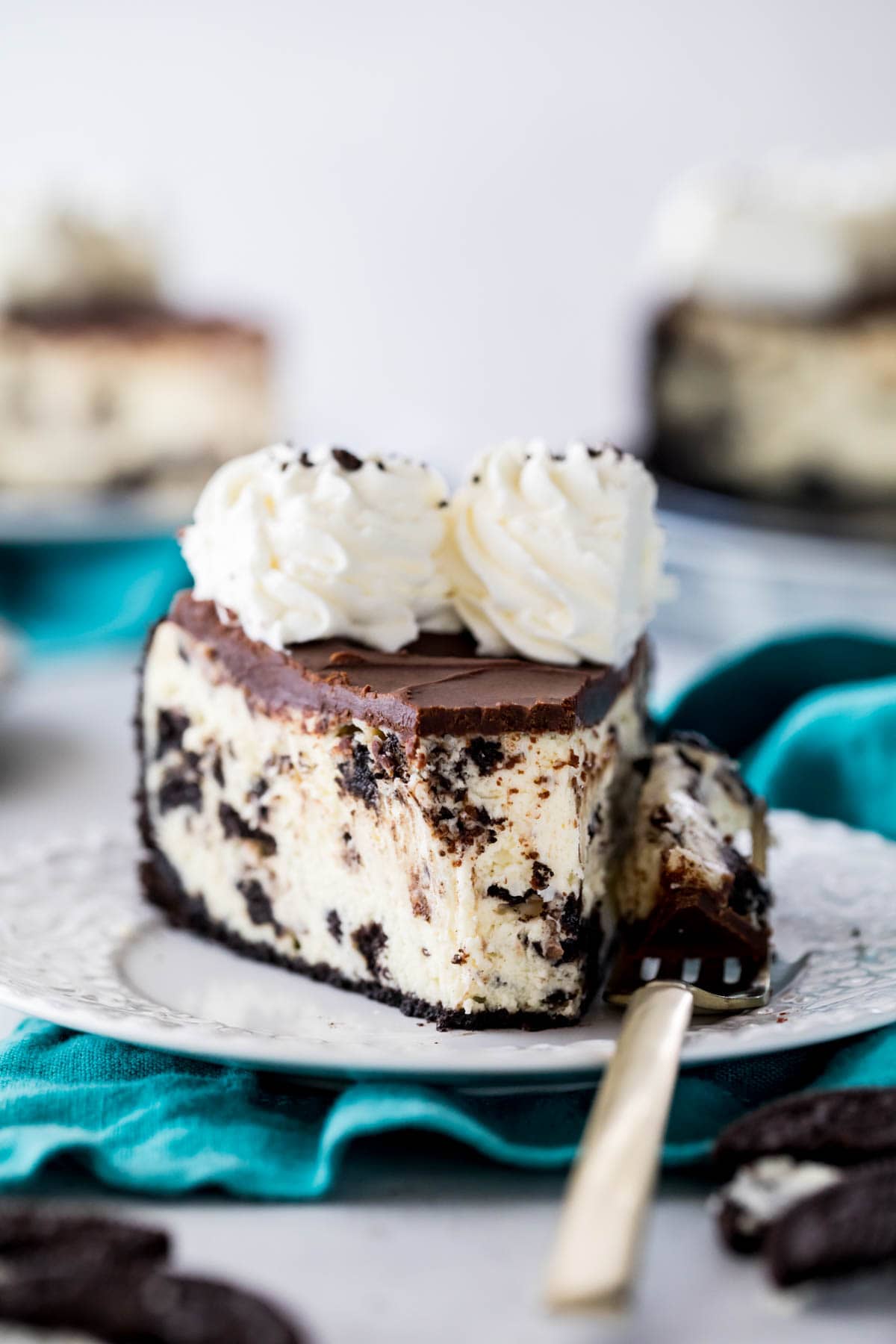 forkful missing from a slice of cheesecake with an oreo crust, crushed oreos in the batter, chocolate ganache top, and whipped cream