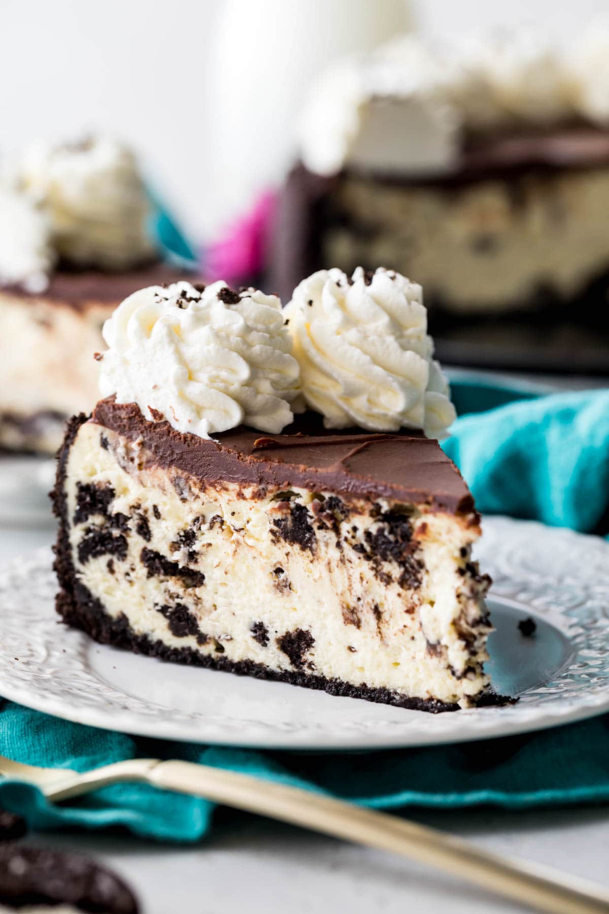 slice of cheesecake with an oreo crust, crushed oreos in the batter, chocolate ganache top, and whipped cream