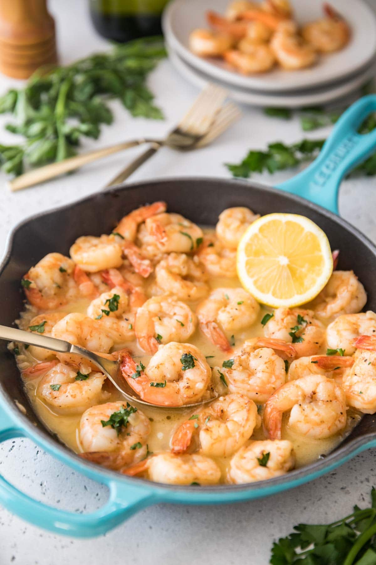 spoon scooping shrimp out of a skillet full of cooked shrimp topped with parsley and lemon