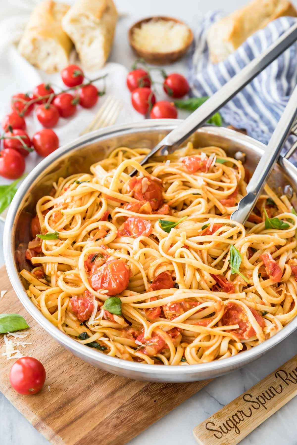 tongs tossing linguine pasta with cherry tomatoes and fresh basil in a stainless steel skillet