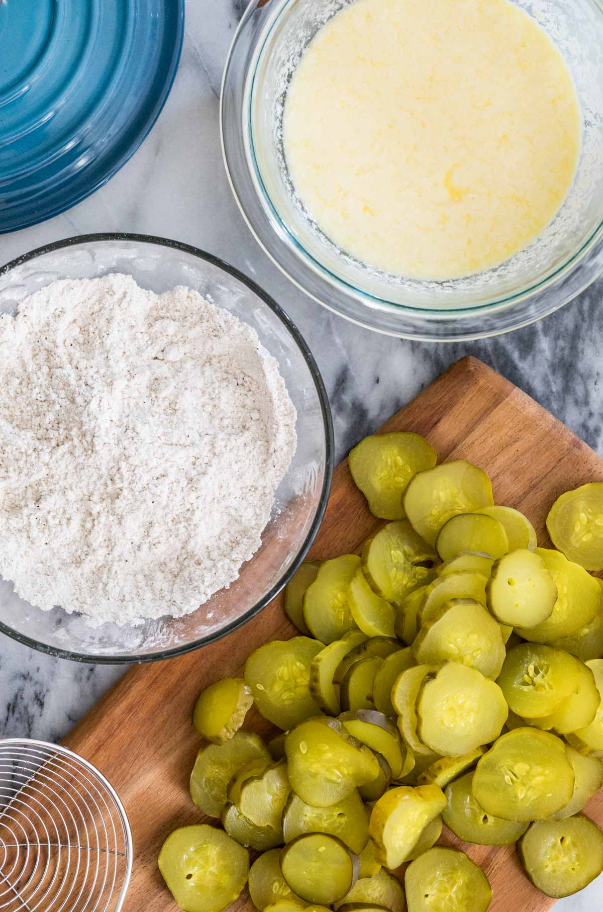 Overhead view of ingredients including pickle slices, a flour mixture, and a buttermilk mixture.