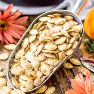 Metal scoop of cinnamon sugar pumpkin seeds surrounded by orange flowers.