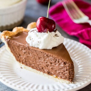 Slice of French silk pie topped with whipped cream and a sweet cherry served on a white plate.