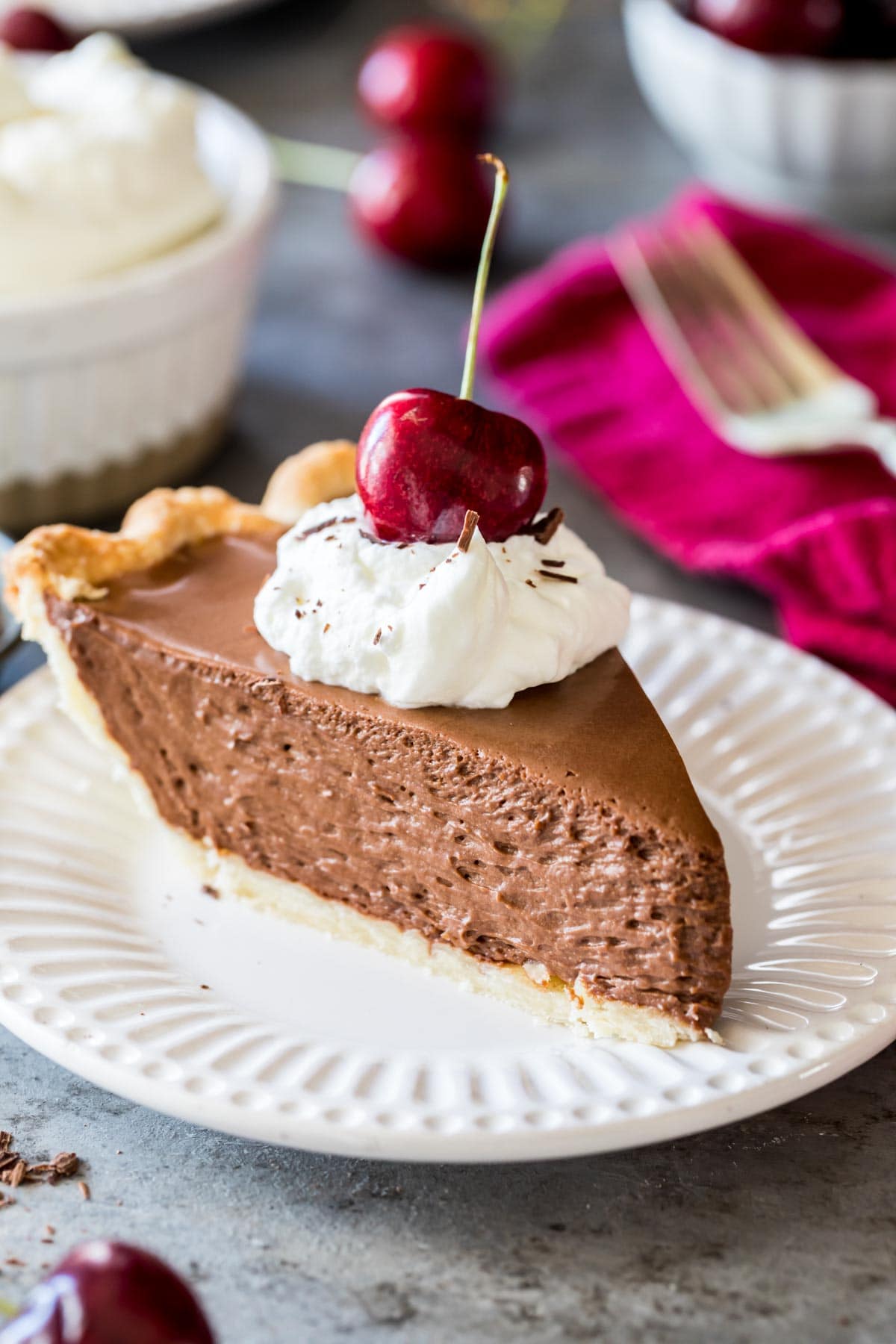 Slice of French silk pie topped with whipped cream and a sweet cherry served on a white plate.