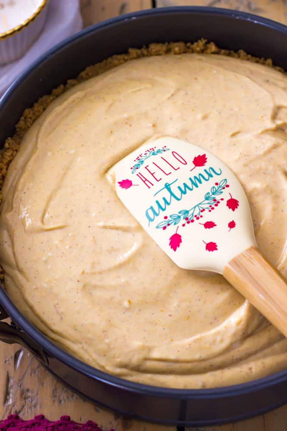 Fall-themed spatula smoothing cheesecake batter after being poured into a springform pan.