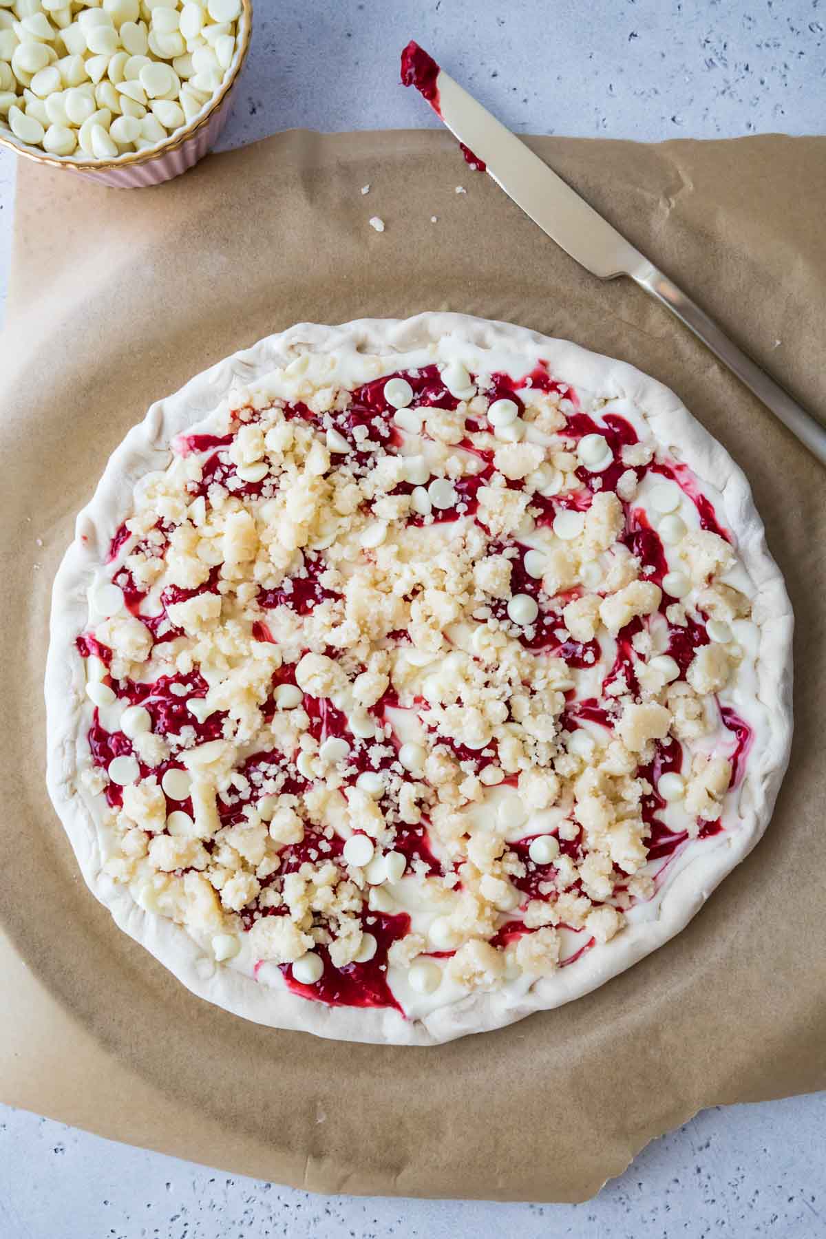 Overhead view of pizza dough topped with cream cheese sauce, raspberry jam, white chocolate chips, and streusel.